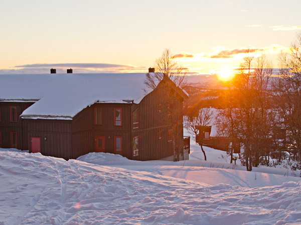 Snødekt fjellhytte med solnedgang i bakgrunnen, omringet av vinterlandskap.