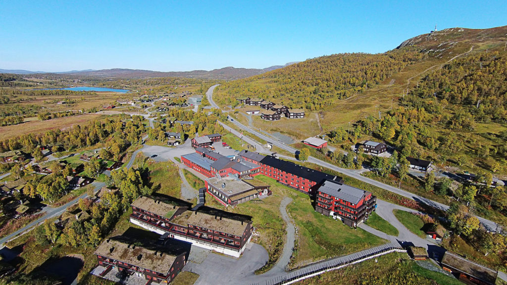 Luftfoto av fjellandskap med røde bygninger, veier og skogkledde åser i vakkert høstvær i Norge.