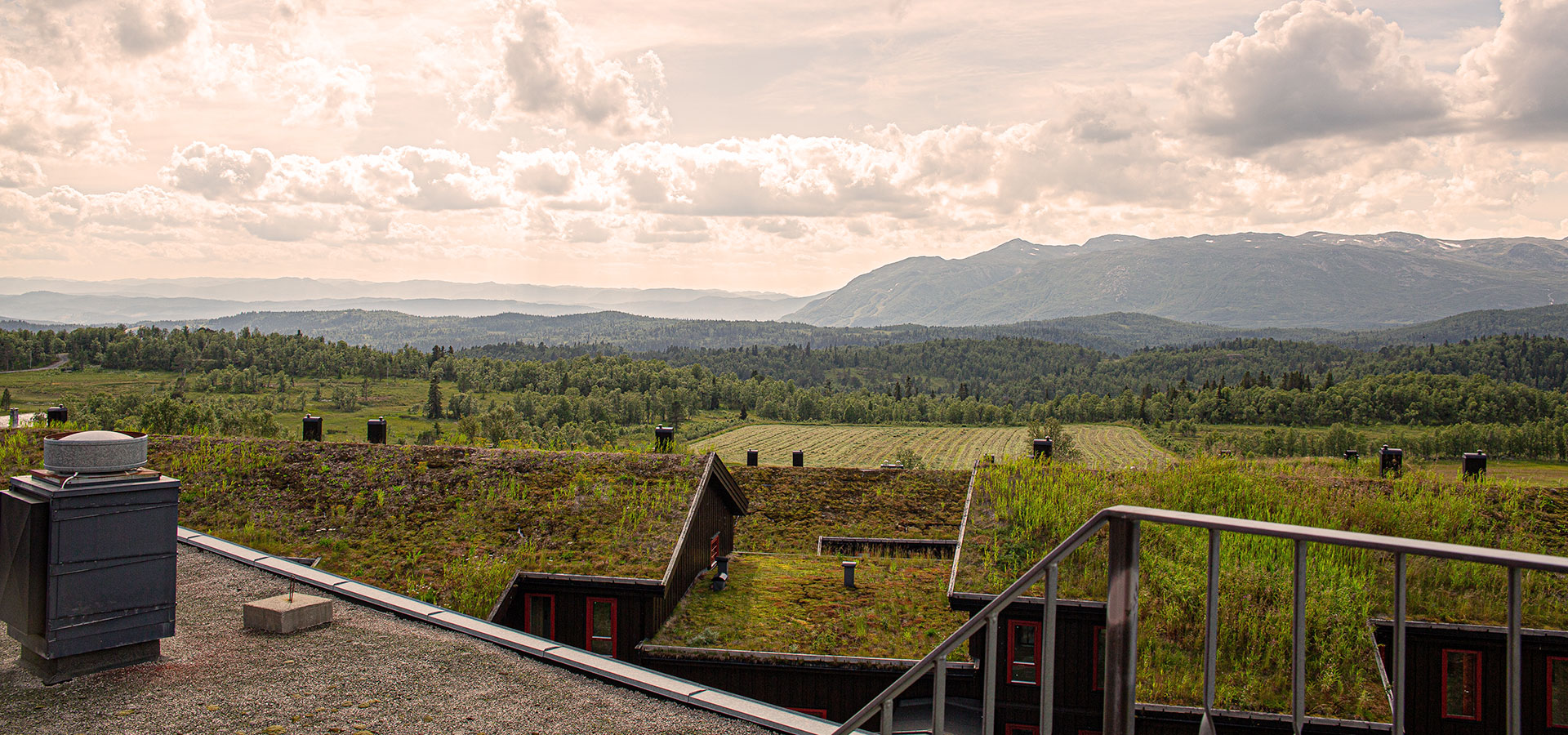 Utsikt over skog og fjell med torvtak i forgrunnen, under delvis skyet himmel, perfekt for turer i norsk natur.