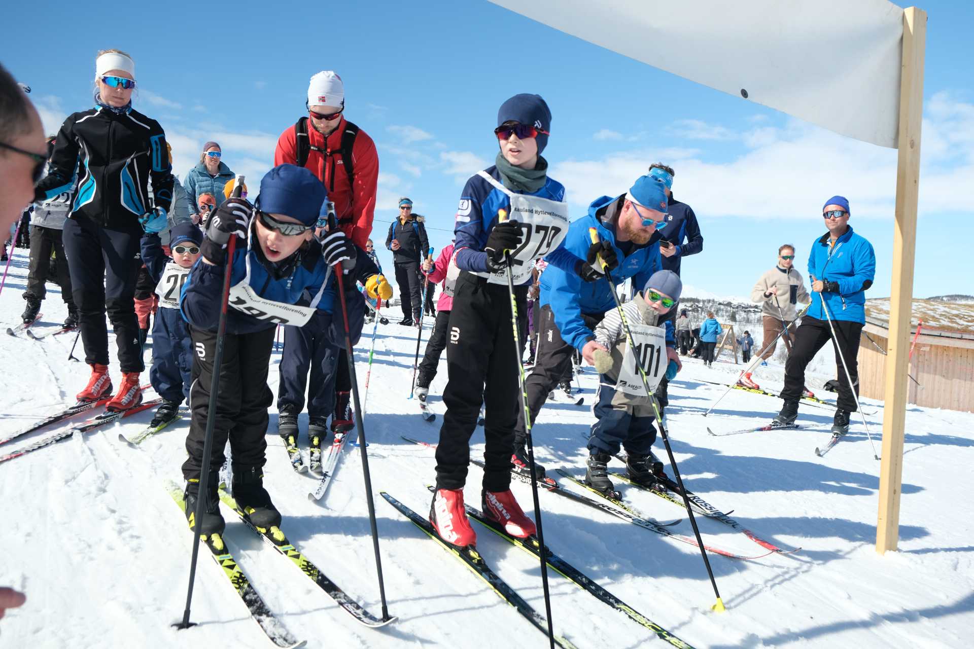 Deltakere i skirenn på snødekket fjell; barn og voksne klare for start i solfylt vær.