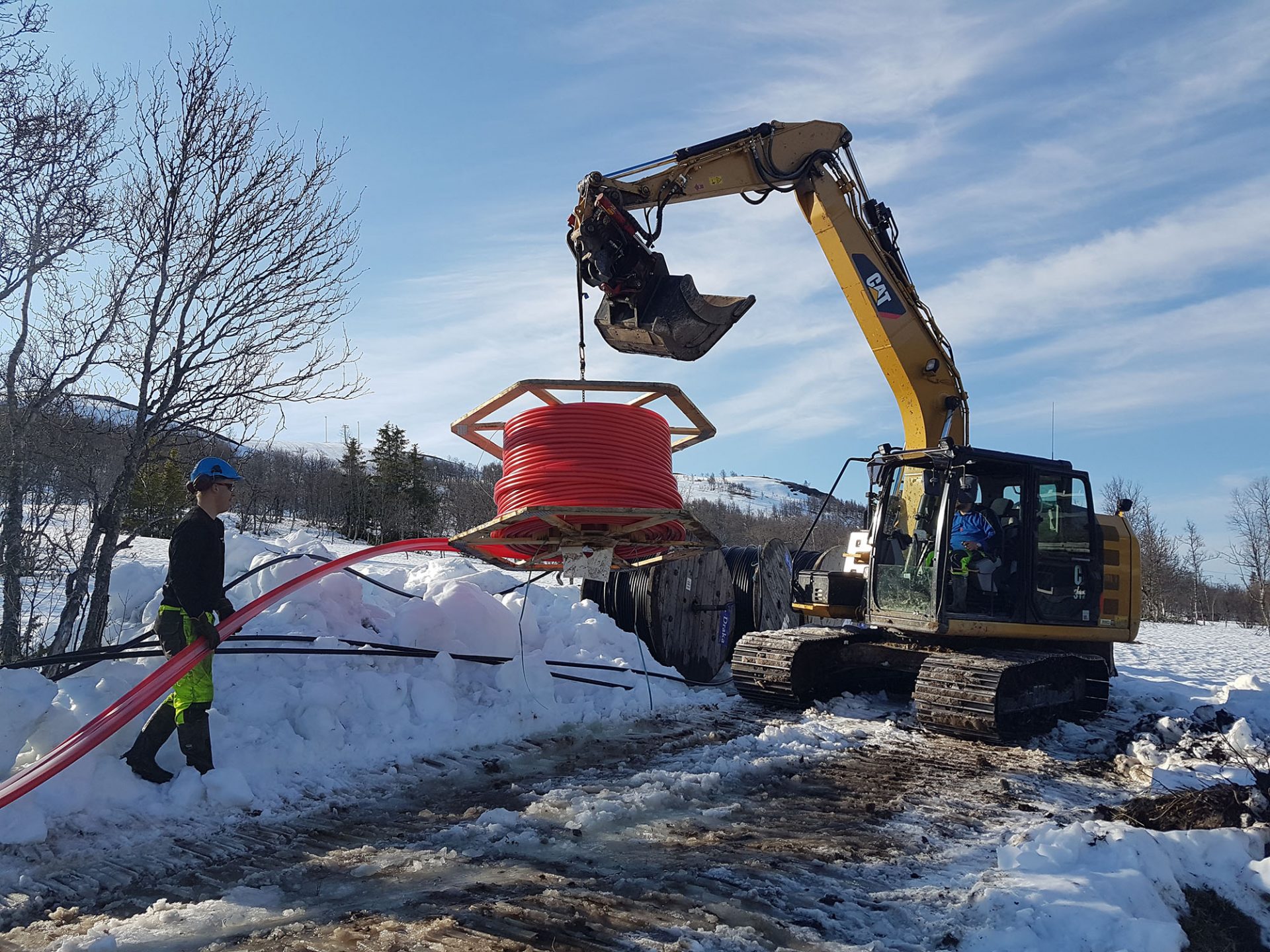 Gravemaskin legger røde kabler i vinterlandskap, arbeidere jobber i snøen.
