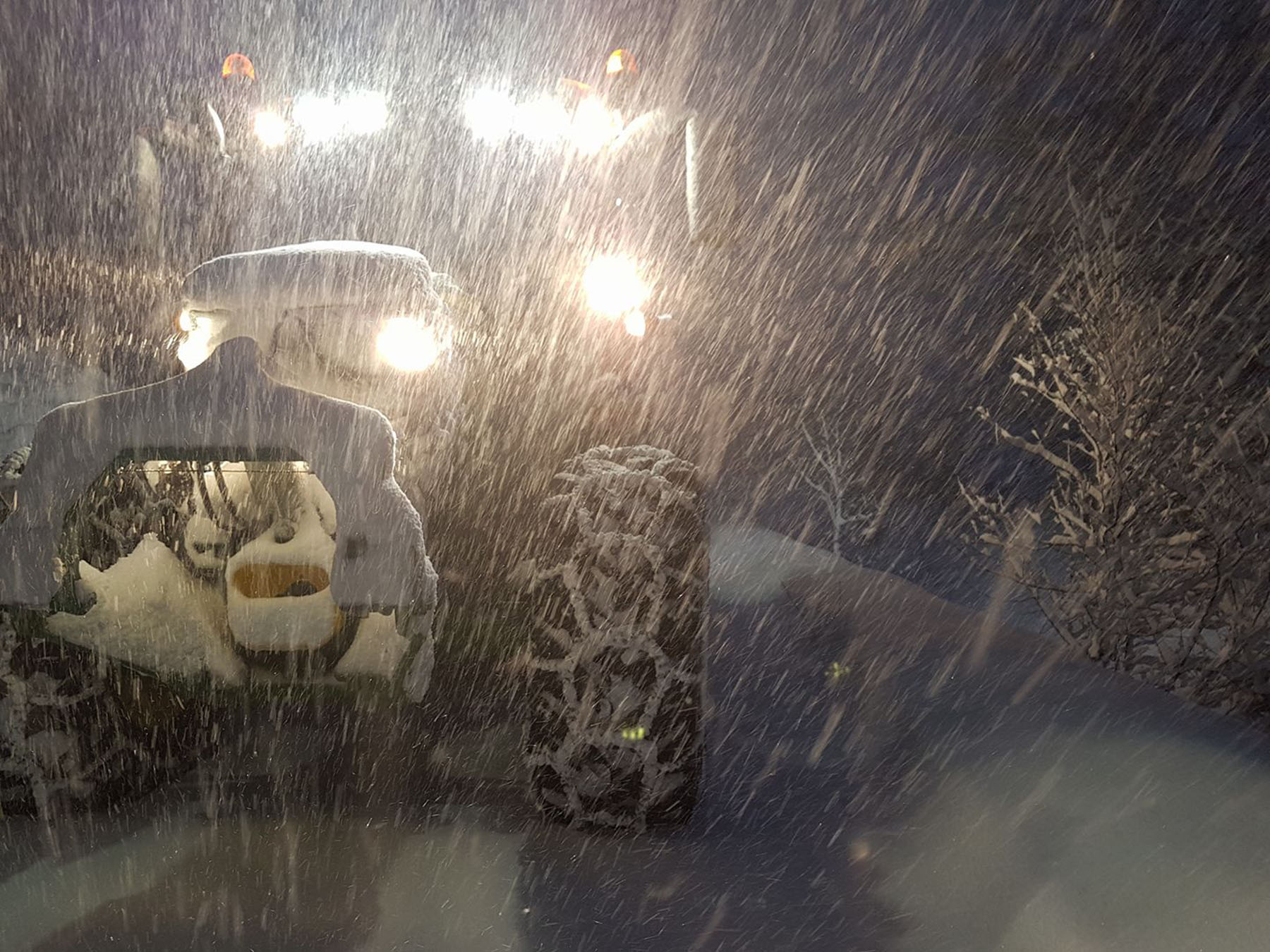 Traktor kjører gjennom kraftig snøstorm i skogen om natten, med frontlysene lysende i mørket.