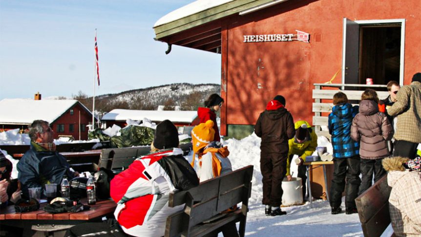 Gruppe mennesker nyter en solrik dag i snøen ved et skihus merket Heishuset med norsk flagg i bakgrunnen.