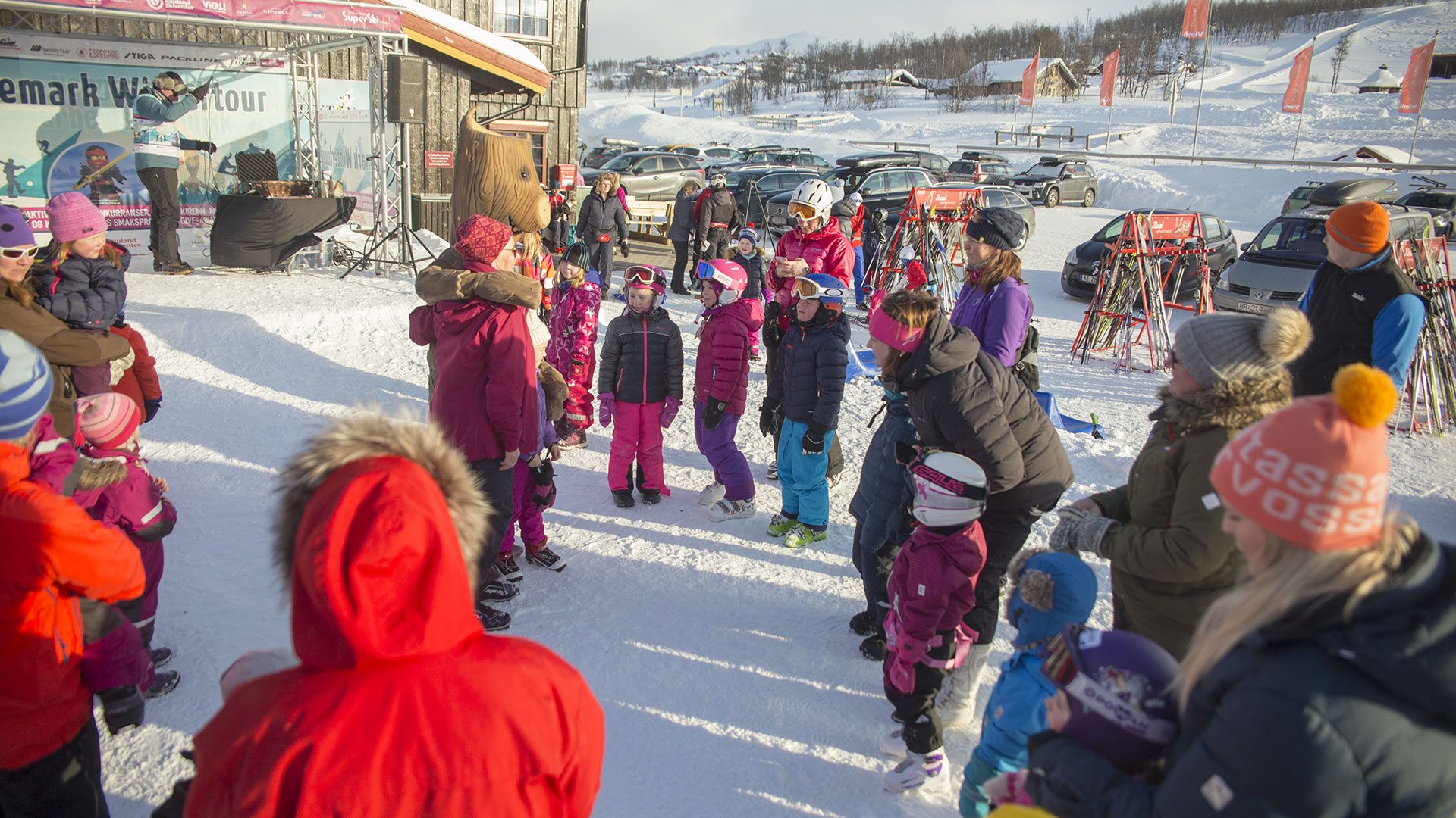 Gruppe av barn og voksne i vinterklær på skisenter, klare for aktiviteter i solrikt vær.