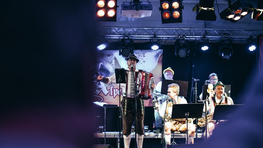 Band spiller på scenen under konsert, musiker med trekkspill og blåseinstrumenter, fargerike lys i bakgrunnen.