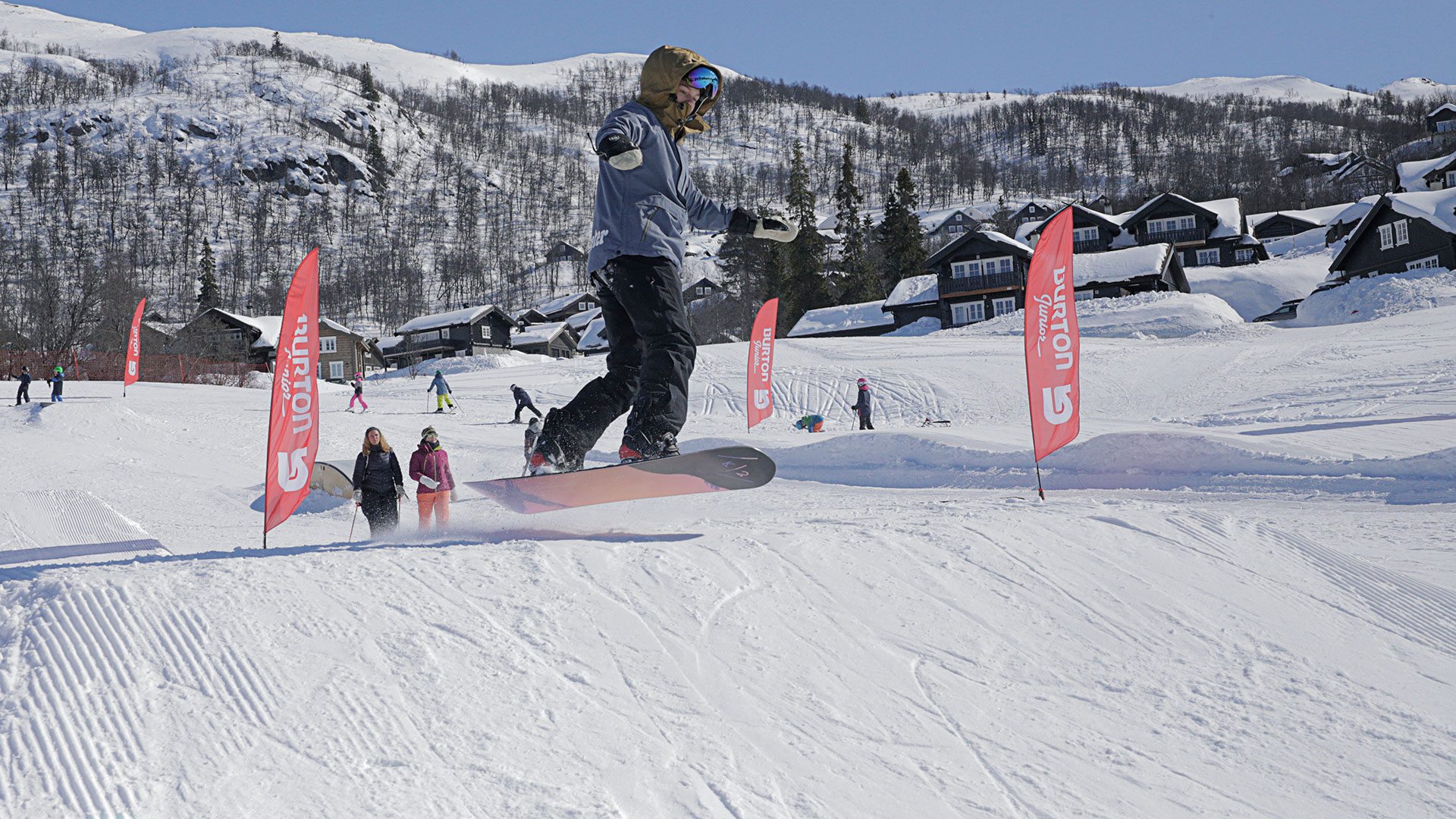 Snowboarder hopper i skibakken, omringet av snødekte fjell og hytter. Klar vinterhimmel i bakgrunnen.