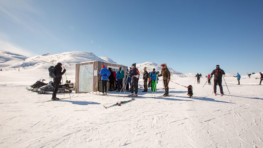 Folk på ski leser kart på fjellet i solskinn, med snødekte topper i bakgrunnen, Norge.
