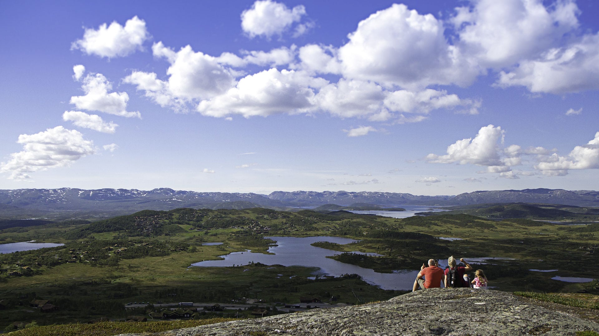 Familie nyter panoramautsikt over norsk fjellandskap med innsjøer og skyer.