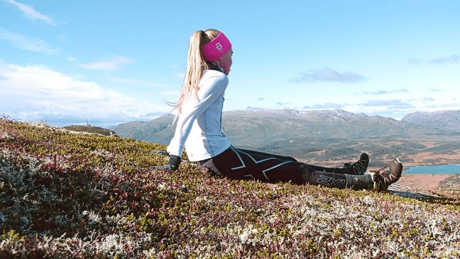 Kvinne hviler i norsk fjellandskap, med blå himmel og fjell i bakgrunnen, iført sporty utstyr og rosa pannebånd.