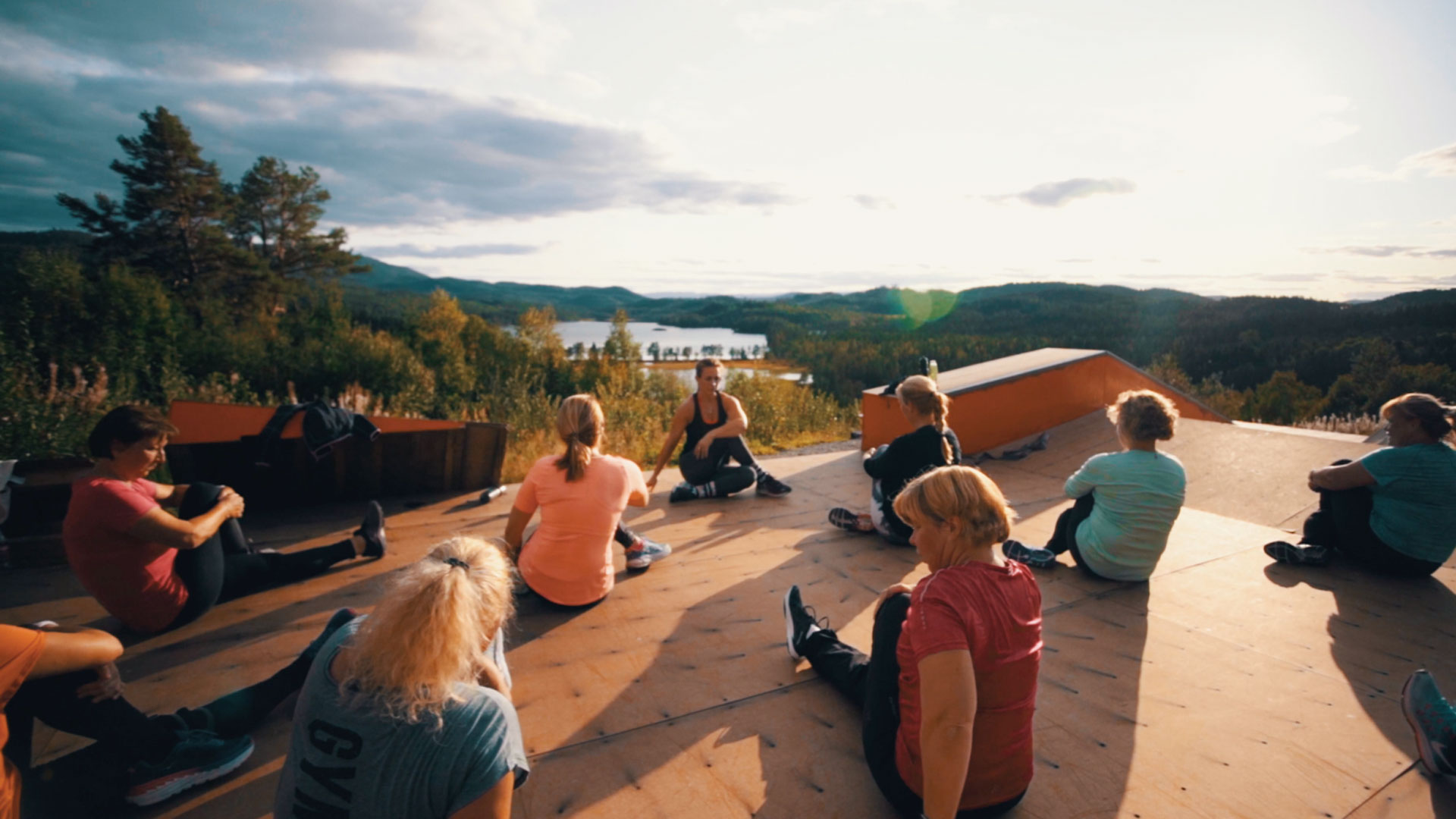 Gruppe yogaøkt i naturskjønn setting med innsjø og skog i bakgrunnen på solfylt dag.