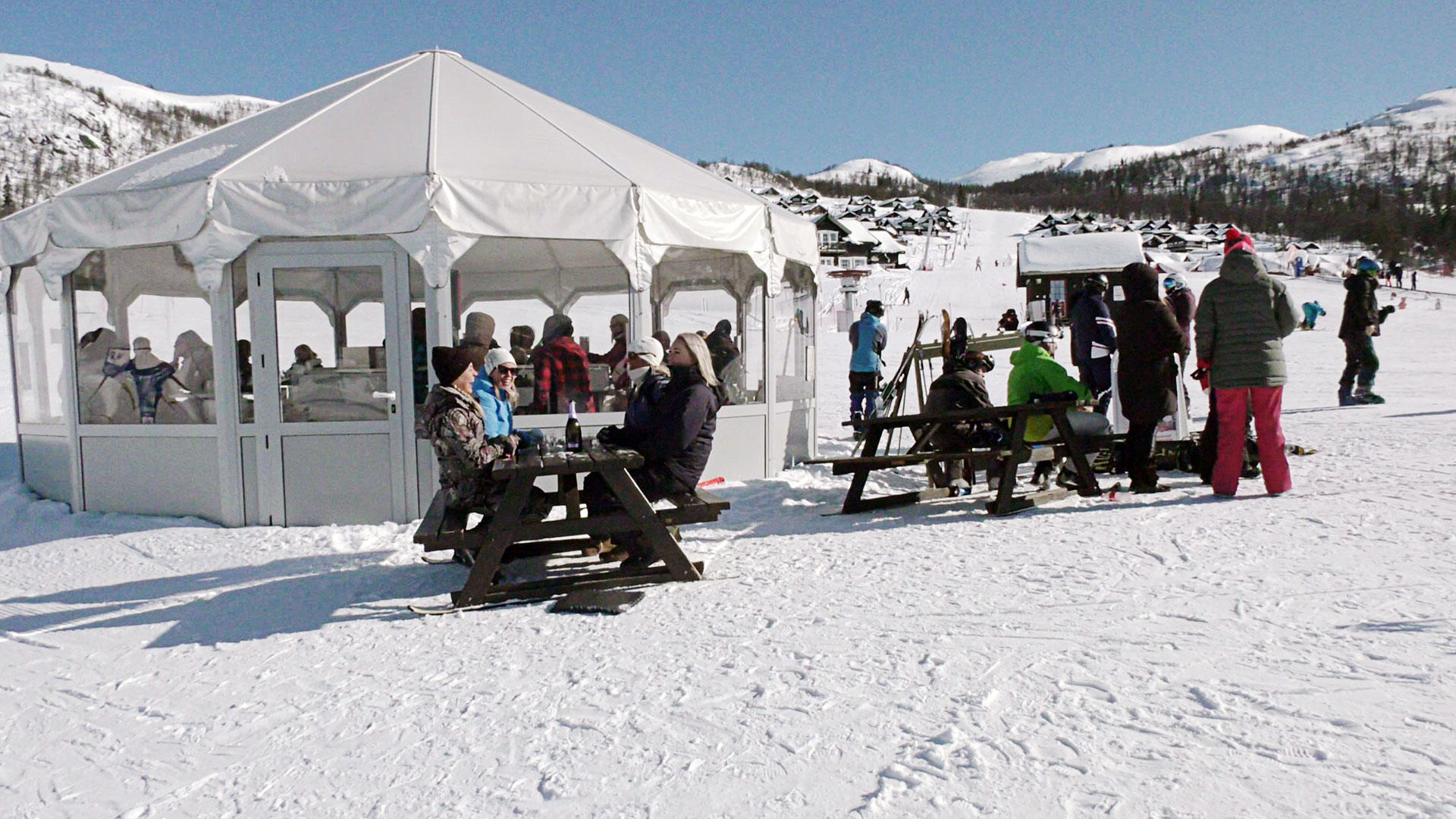Folk nyter sol og snø på en uteplass ved skianlegg, snødekte fjell i bakgrunnen.