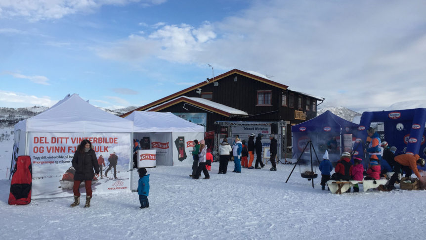 Vinterarrangement ved skianlegg med telt og folk som deltar i aktiviteter. Snødekte fjell i bakgrunnen.