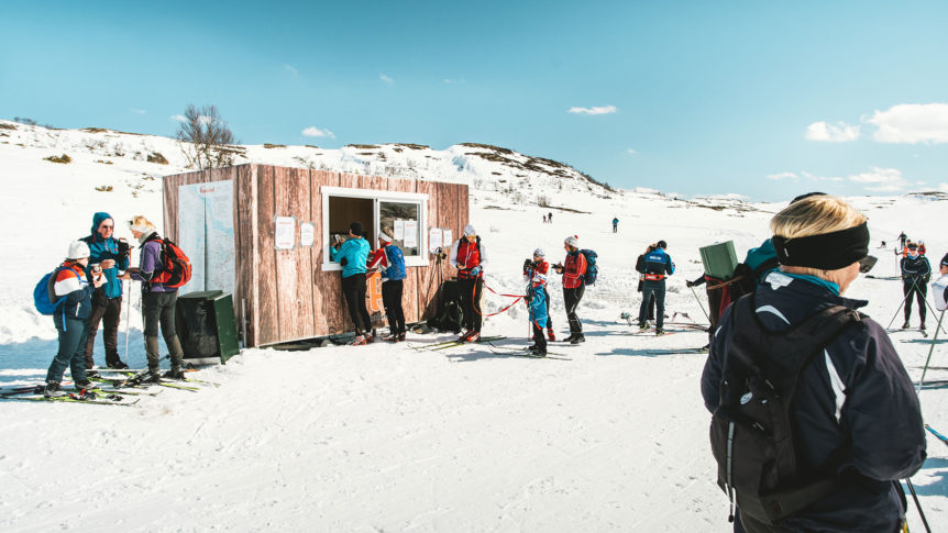 Langrennsløpere samles ved en kiosk i snødekt fjellandskap under klar blå himmel.