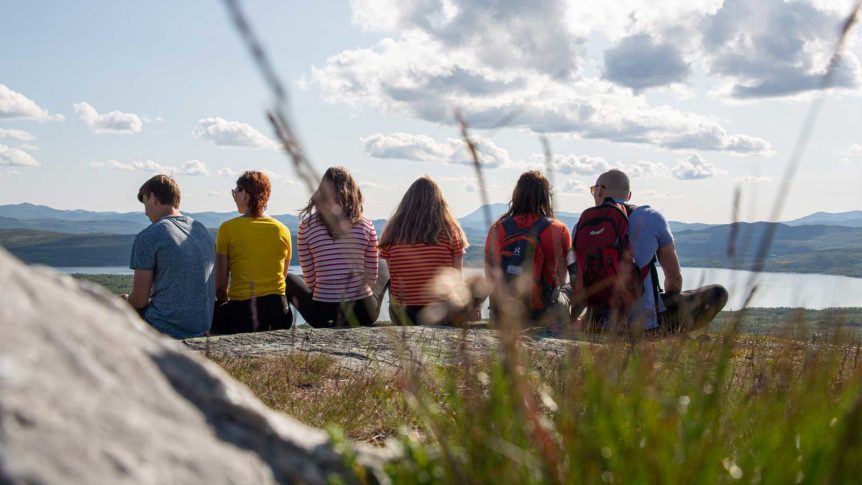 Seks personer nyter utsikten fra en fjelltopp med innsjø og fjell i horisonten under en klar himmel.