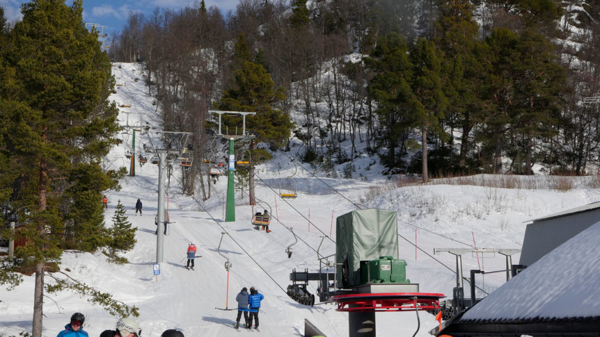 Skitrekk i snødekte omgivelser med folk og skog i bakgrunnen, solfylt vinterdag i skisenter.
