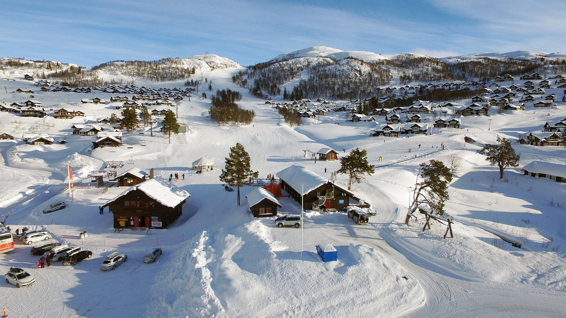 Vinterlandskap med snødekte fjell, hytter, skiløpere og biler på en solfylt dag i Norge.
