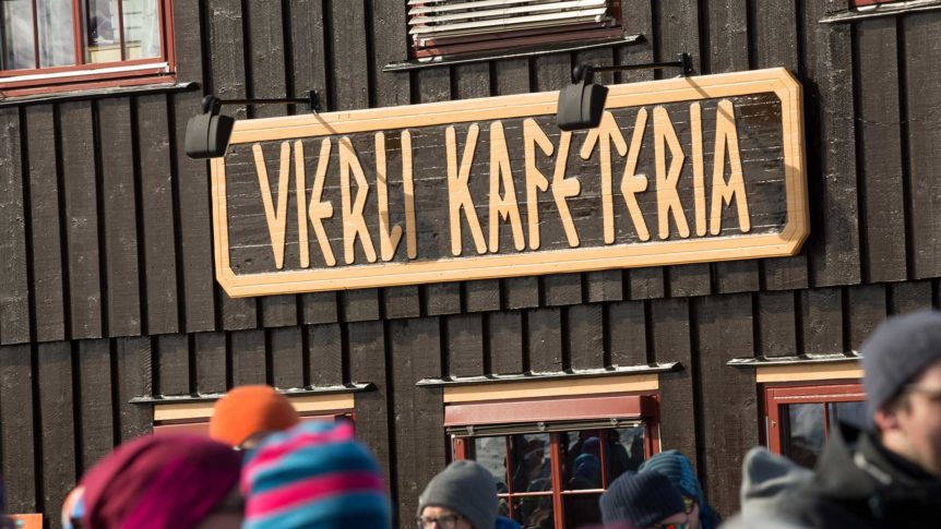 Skilt for Vekli Kafeteria på en brun trebygning, med flere mennesker foran.