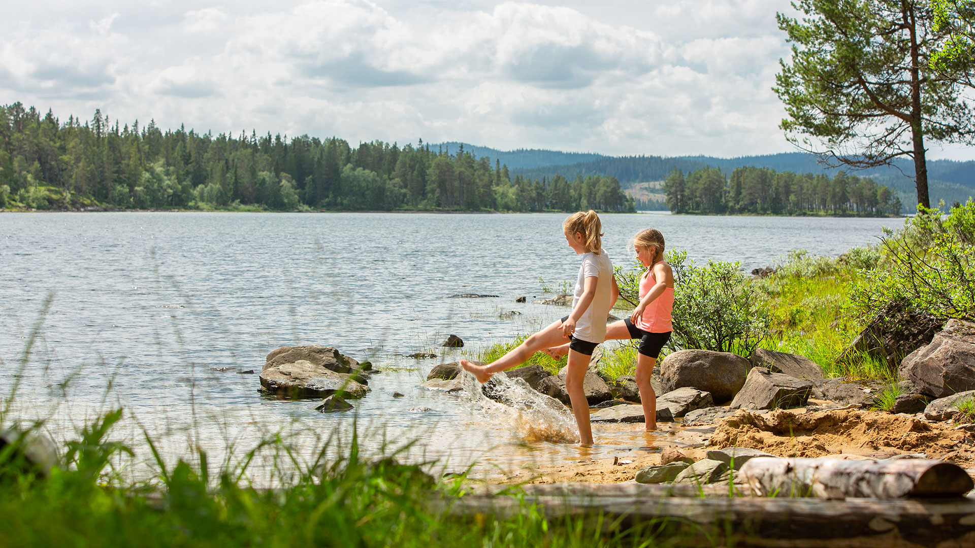 To barn leker ved innsjøen på en solfylt dag omgitt av skog i Norge.