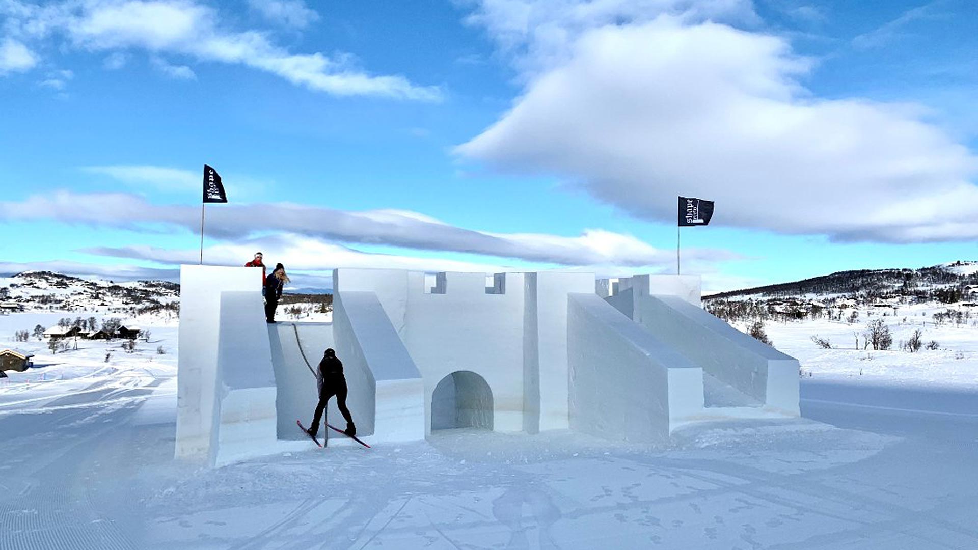Snøslott med mennesker på ski ved solsydde fjell under blå himmel og flagg i Geilo, Norge.