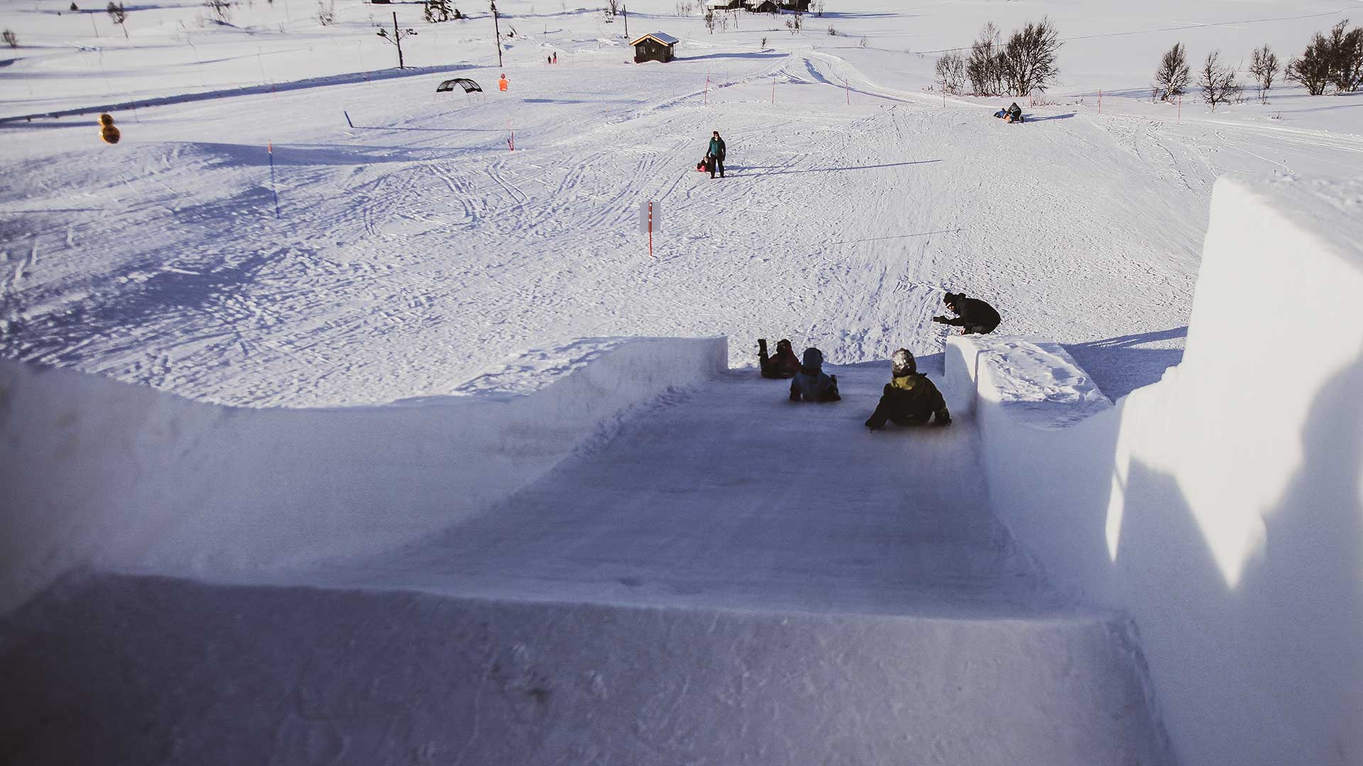 Barn leker på stor snøsklie i vintersportsområde. Snødekt landskap i bakgrunnen.