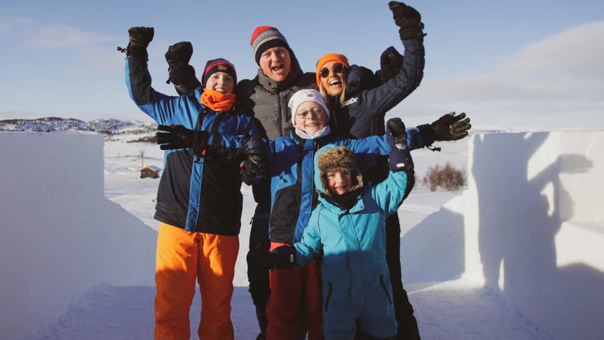 Gruppe glade mennesker i vinterklær jubler på snøfylt landskap under blå himmel.