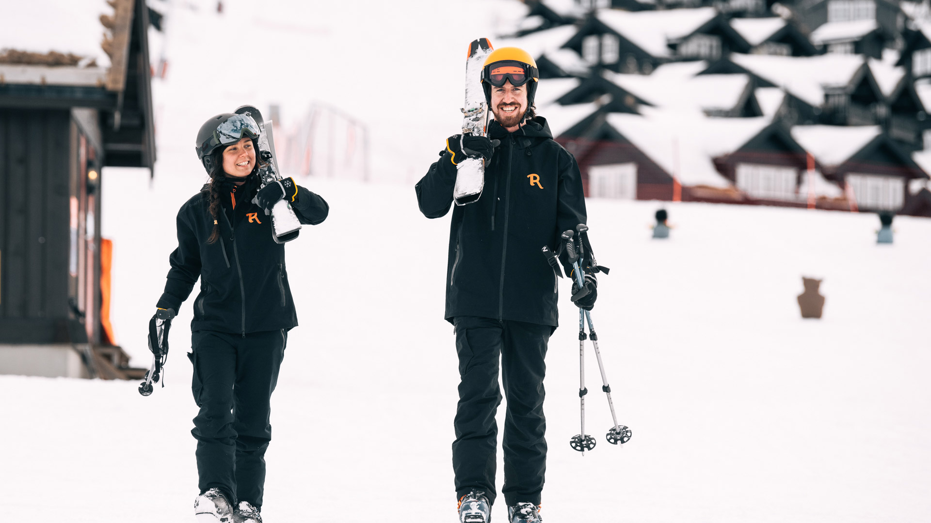 To personer i skiutstyr smiler og går på snødekket bakke foran skihytter.