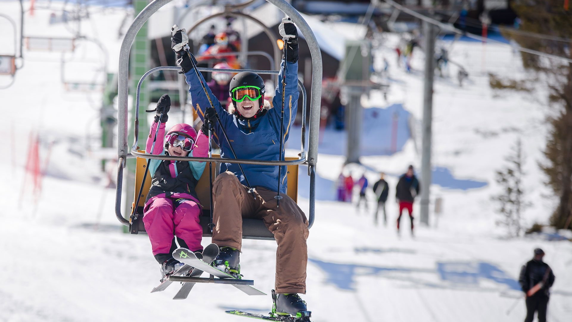 To personer på skiheis, smilende med skiutstyr på, i et snødekt skianlegg.