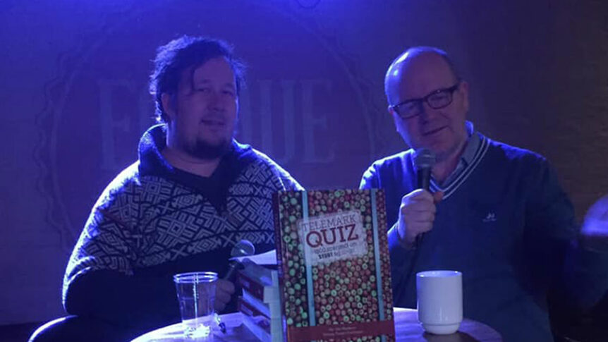 To personer med mikrofoner deltar på quizkveld, en bok om Telemark Quiz på bordet foran dem.