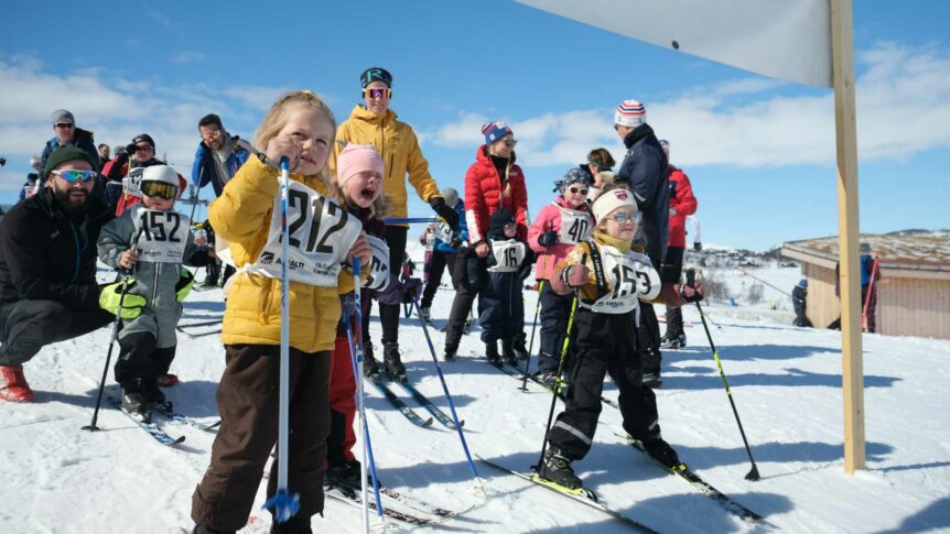 Barn og voksne klare for skirenn i solfylt vinterlandskap. Nummererte startnumre på deltakerne.
