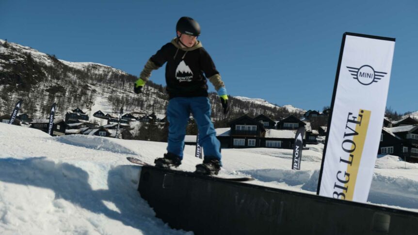 Ung snowboarder balanserer på rail i skiområde, omgitt av snødekte fjell og hytter under blå himmel.