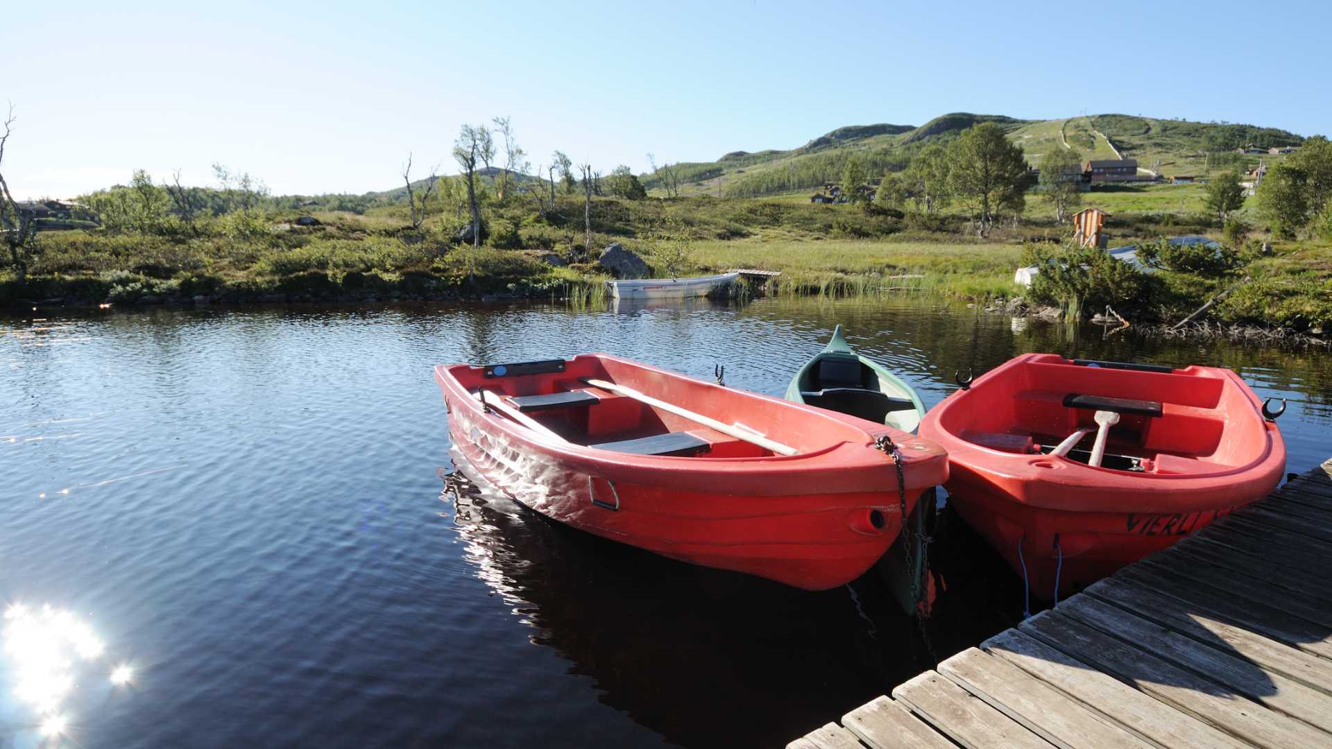 Røde båter ved bryggen på en innsjø om sommeren, omgitt av norsk natur og grønne åser.