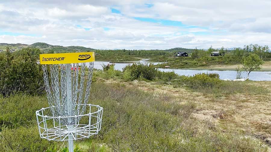 Frisbeegolfkurv ved siden av innsjø i naturskjønt landskap med hytter i bakgrunnen, solrik dag.