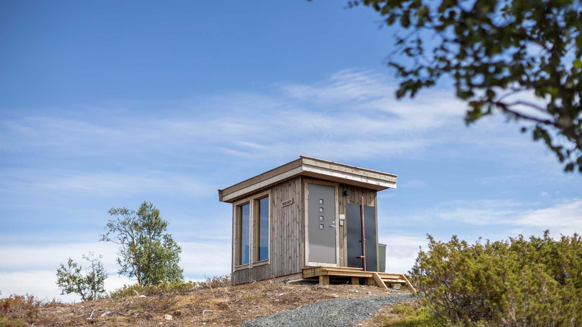 Liten hytte på en ås med blå himmel og trær, perfekt for rolige utflukter i norsk natur.