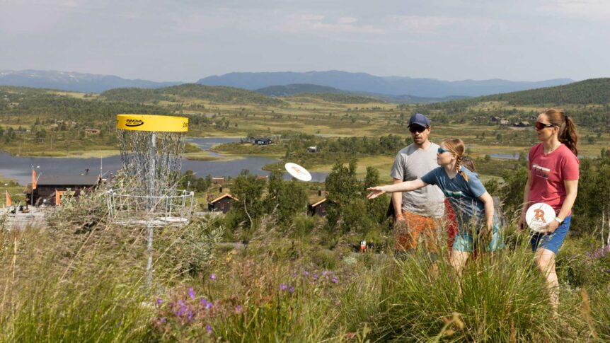 Familie spiller frisbeegolf i norsk landskap med innsjø og fjell i bakgrunnen.