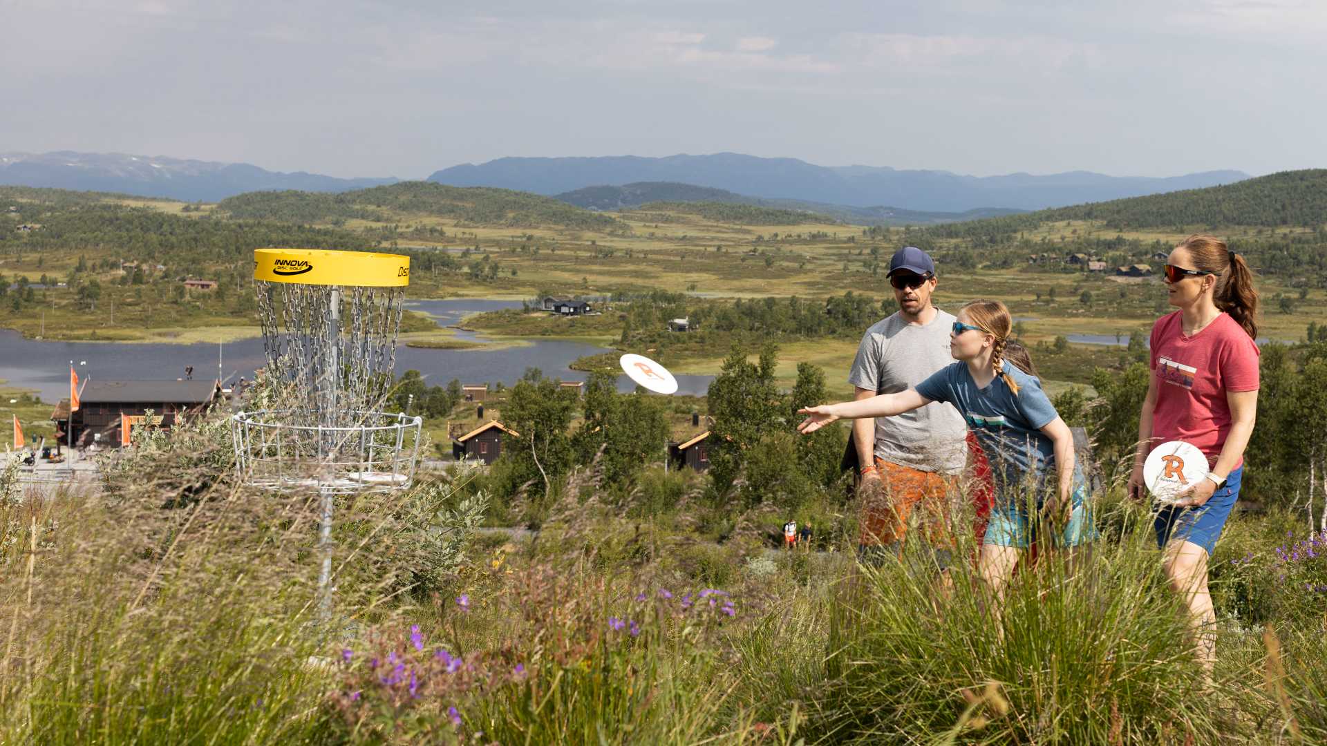 Familie spiller frisbeegolf i norsk landskap med innsjø og fjell i bakgrunnen.