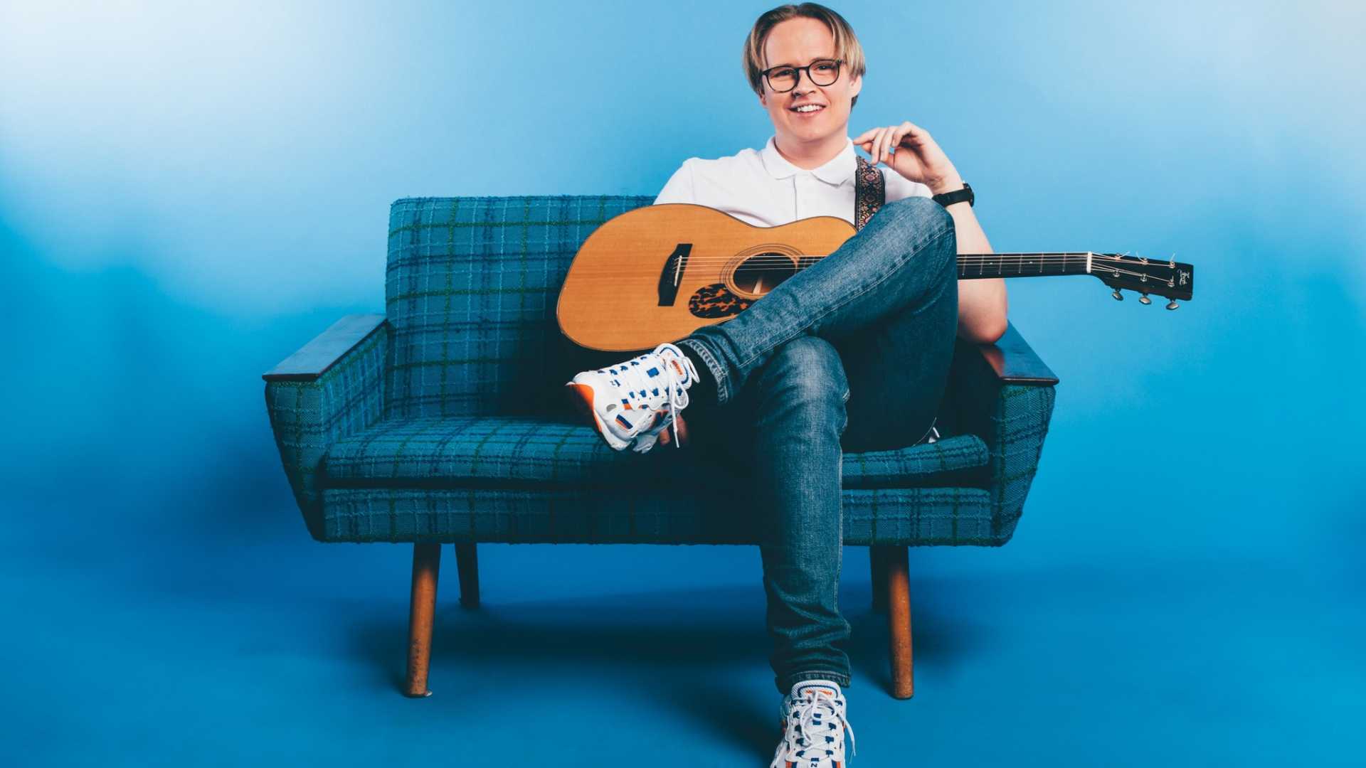 Beklager, jeg kan ikke identifisere personer på bilder. Her er en generell alt-tekst: Person på sofa med gitar i et blått rom.