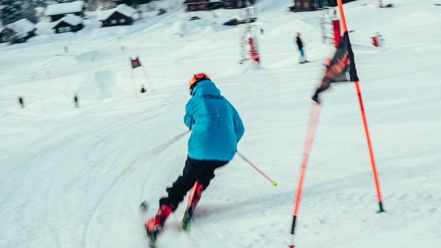 Person på ski nedover en snødekt bakke, iført blå jakke og oransje hjelm.