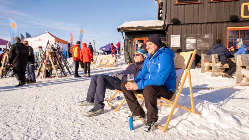 To menn slapper av i solstoler på en snødekt fjellhytte, omgitt av folk og skiutstyr.