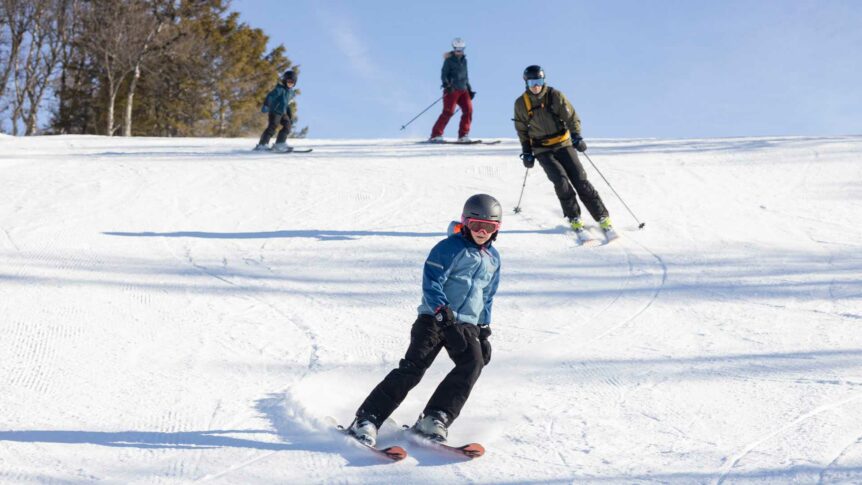 Personer på ski ned en solfylt bakke om vinteren, omgitt av snødekte trær.