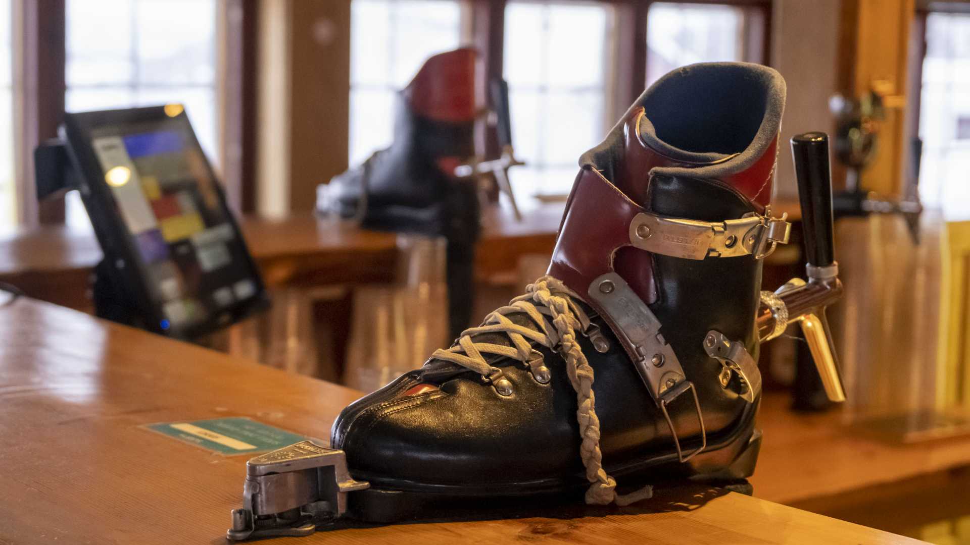 Skiboot-basert ølkran på en trebenk i en rustikk bar med et kassaapparat i bakgrunnen.