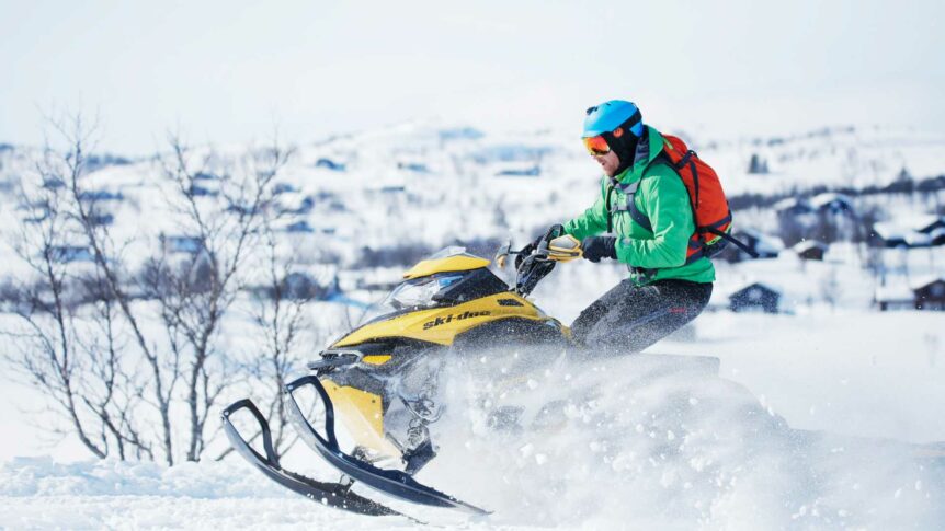 En person på snøscooter kjører gjennom snølandskap iført grønn jakke og blå hjelm, vinteraktiviteter i Norge.
