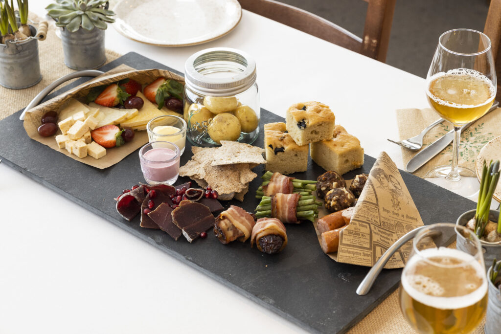 Planke med assorterte småretter og øl: ost, frukt, poteter, focaccia, spekemat og grønnsaker, klar til servering.