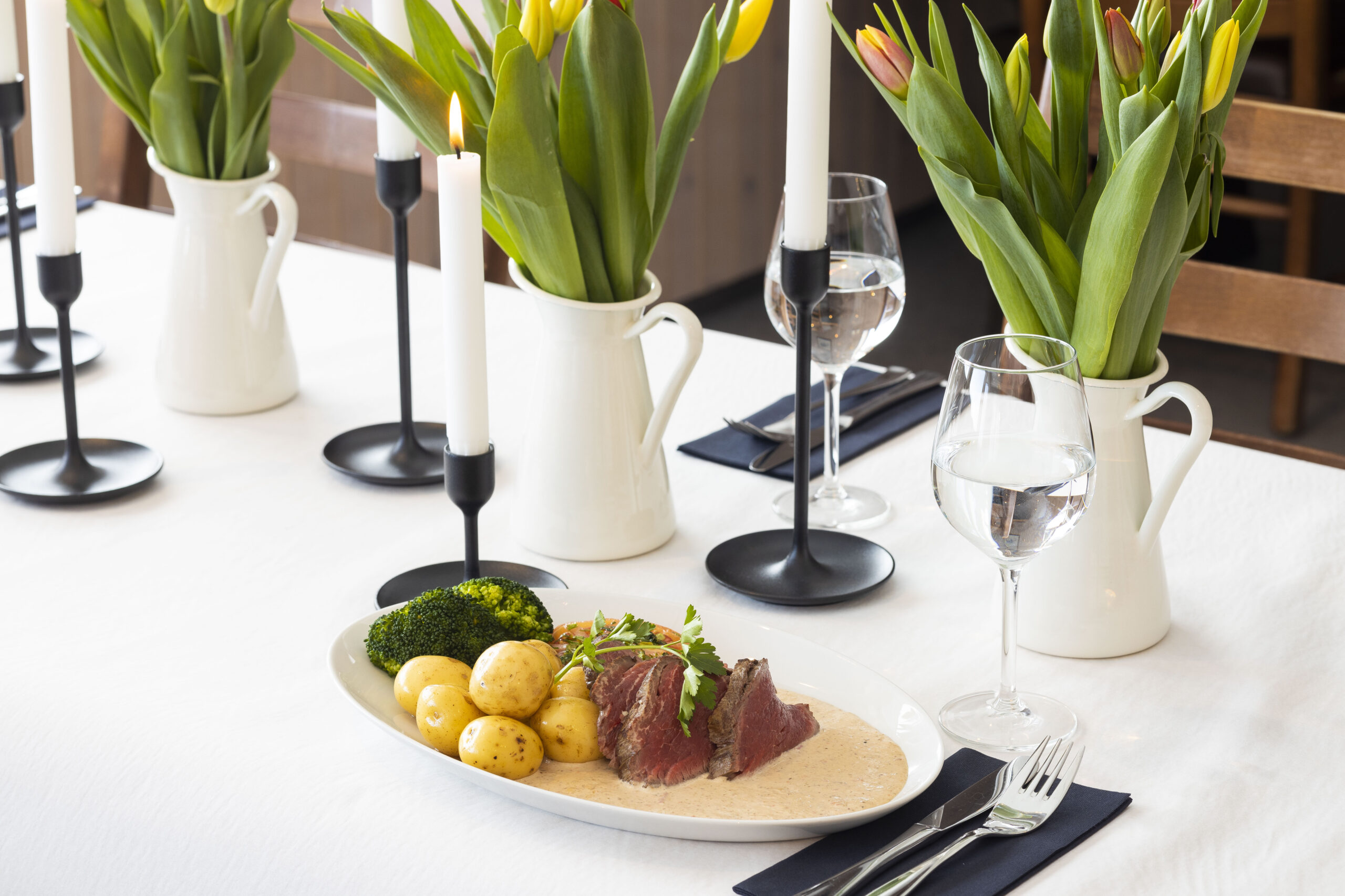 Bord med festmåltid: Ovnsstekt kjøtt, poteter, saus, brokkoli, tulipaner, stearinlys og vinglass i elegant setting.