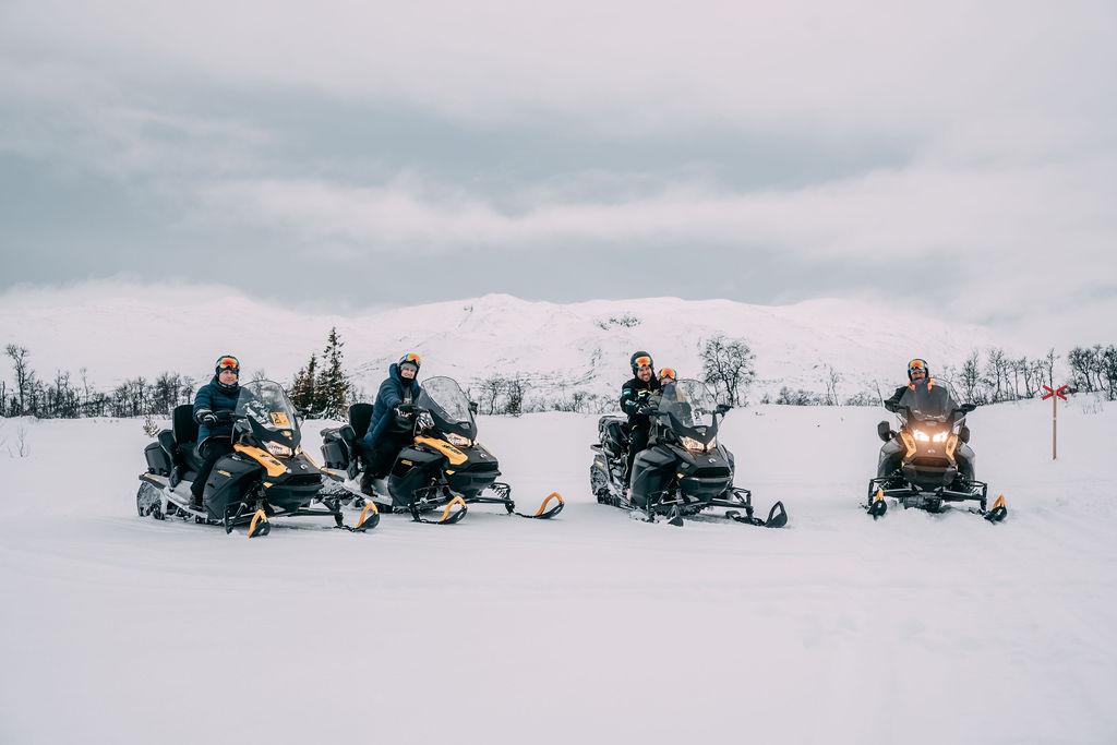 Fire personer på snøscootere i et vinterlandskap med fjell i bakgrunnen.