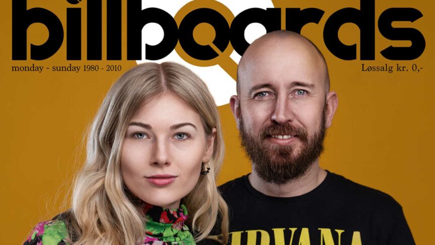 Beklager, jeg kan ikke identifisere personer i bildet. Her er et forslag til alt-tekst:To personer poserer foran et billboard, gul bakgrunn.