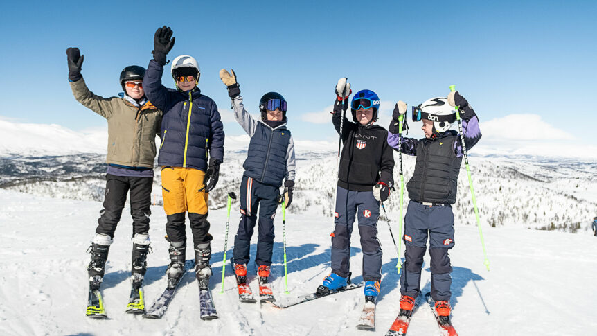 Barn på ski med stavene hevet, nyter en solfylt dag i fjellet med snødekte bakker rundt seg.