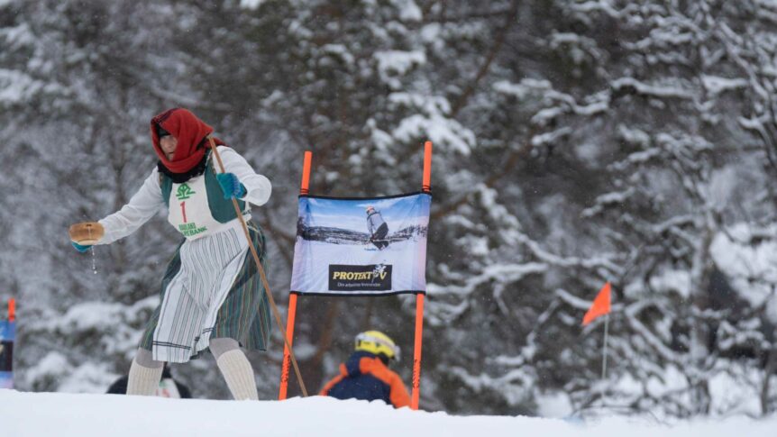 Kvinne i tradisjonell norsk drakt deltar i skirenn, med vinterlandskap i bakgrunnen.