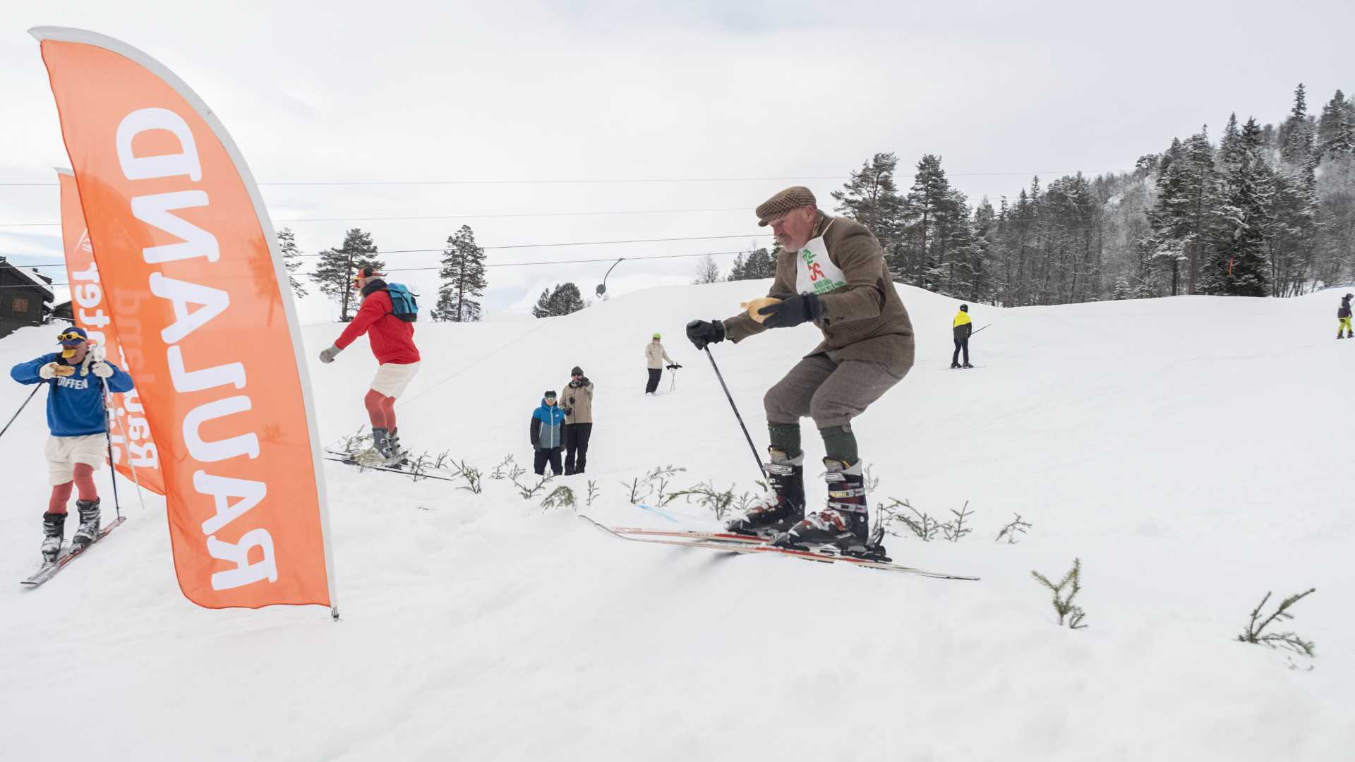 Skikonkurranse i snødekte fjell, folk i ulike antrekk står på ski med bakgrunn av trær og en Røros-vimpel.