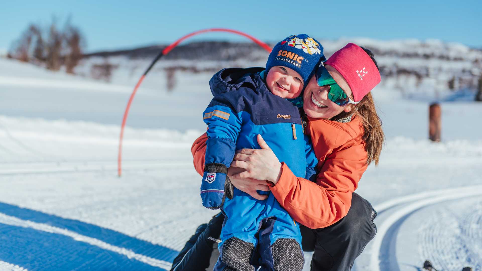 Smilende voksen og barn koser seg i vinterlandskap på skitur under solfylt himmel.