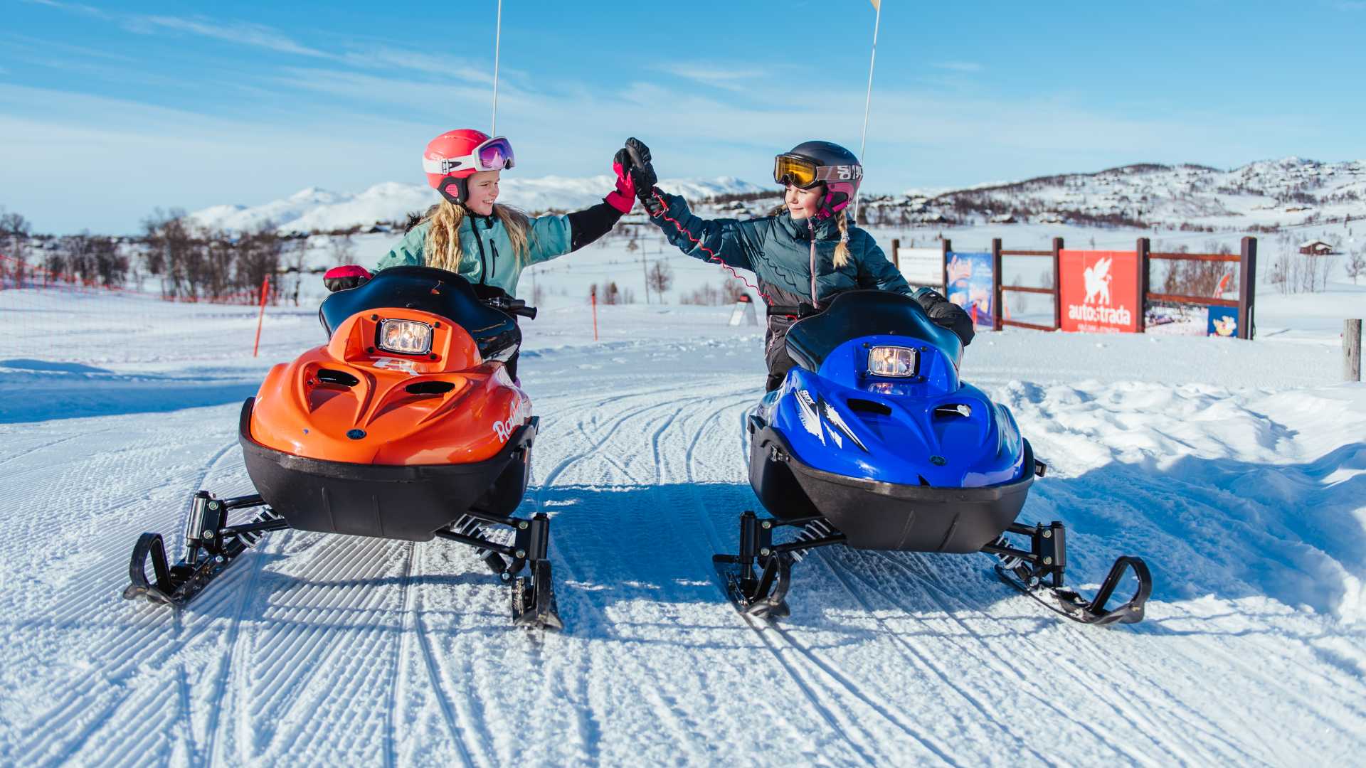 To personer på snøscootere high-fiver i solfylt vinterlandskap med blå himmel i bakgrunnen.